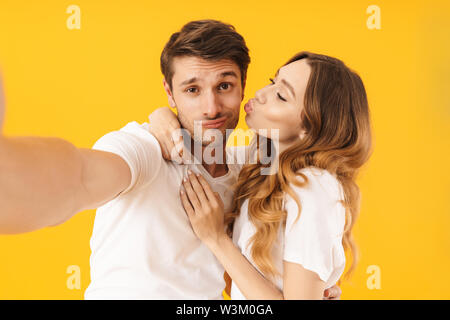 Ritratto di uomo felice guardando la fotocamera e tenendo selfie foto mentre adorabile ragazza baciare lui sulla guancia isolate su sfondo giallo Foto Stock