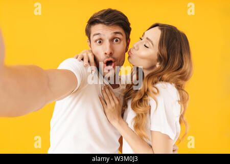 Ritratto di attraente uomo che guarda la fotocamera e tenendo selfie foto mentre adorabile ragazza baciare lui sulla guancia isolate su sfondo giallo Foto Stock