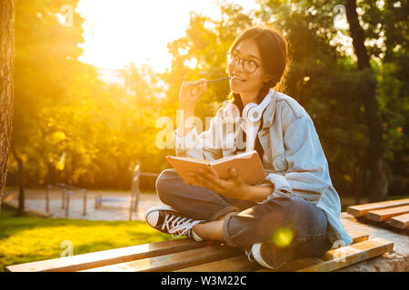 Immagine di un pensiero sognando il giovane studente ragazza indossando occhiali seduti all'aperto nel parco naturale di ascolto di musica con le cuffie la scrittura di note. Foto Stock