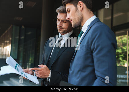Immagine di uomini d'affari di successo partner vestito in abito formale in piedi al di fuori centro uffici e trattenere i grafici della carta durante la riunione di lavoro Foto Stock