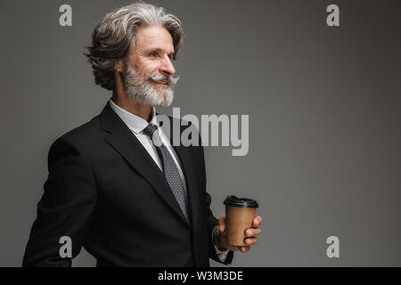 Immagine di mezza età professore uomo formale da indossare tuta nera e sorridente holding bicchiere di carta isolate su uno sfondo grigio Foto Stock