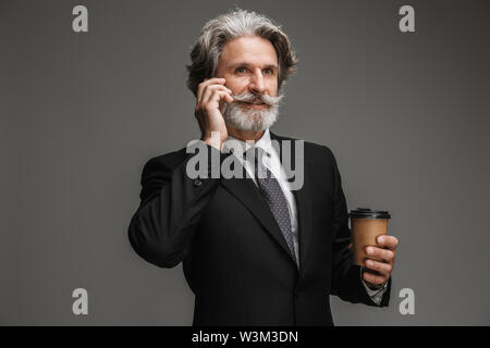 Immagine di gioiosa imprenditore adulti formale da indossare tuta nera tenendo bicchiere di carta e parlare sullo smartphone isolate su uno sfondo grigio Foto Stock
