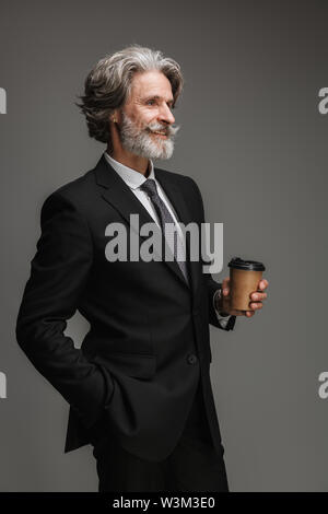 Immagine del professore adulto uomo formale da indossare tuta nera e sorridente holding bicchiere di carta isolate su uno sfondo grigio Foto Stock