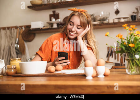 Foto di un piacere giovane ragazza bionda chef in cucina La cucina utilizzando il telefono cellulare. Foto Stock