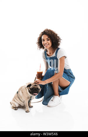 Immagine di adorabili americano africano donna che indossa una tuta in jeans tenendo una tazza di plastica mentre posa con la pug isolate su sfondo bianco Foto Stock
