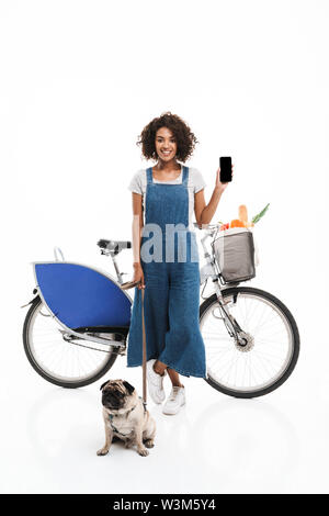 Immagine di bruna americano africano donna smartphone di contenimento mentre in piedi con la sua pug e bicicletta isolate su sfondo bianco Foto Stock