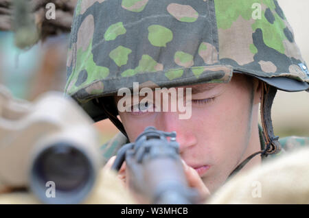 Giovane maschio Seconda Guerra Mondiale ri-enactor raffiguranti Esercito Tedesco soldato guardando giù il sito di un fucile direttamente alla fotocamera. Contatto visivo Foto Stock