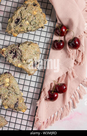 Close up ciliegia e cioccolato bianco scones su una cottura nero rack e marmo rosa sfondo con un tovagliolo e verticale delle ciliegie Foto Stock