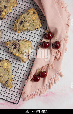 Ciliegia e cioccolato bianco scones su una cottura nero rack e marmo rosa sfondo con un tovagliolo e verticale delle ciliegie Foto Stock