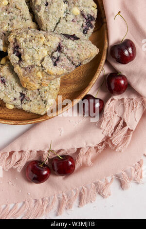 Ciliegia e cioccolato bianco scones su una lastra di legno con un tovagliolo rosa e verticale delle ciliegie Foto Stock