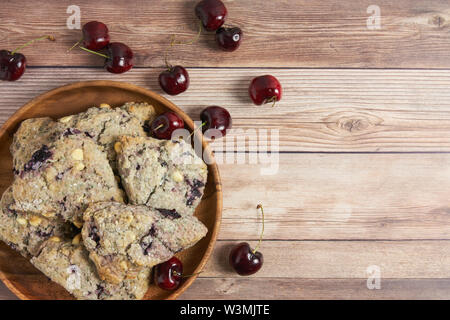 Piastra in legno di ciliegio e il cioccolato bianco scones su un tavolo di legno sfondo con ciliegie fresche Foto Stock