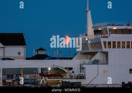 Eclissi lunare parziale, Southampton, Hampshire, Inghilterra, Regno Unito, 16th luglio 2019. Un'eclissi lunare parziale e un cielo nebuloso all'orizzonte fanno diventare la luna arancione rosso durante l'alba serale sulla costa meridionale. In primo piano, il traghetto Red Falcon Funnel dell'Isola di Wight è appena attraccato nel porto per lo sbarco. Foto Stock