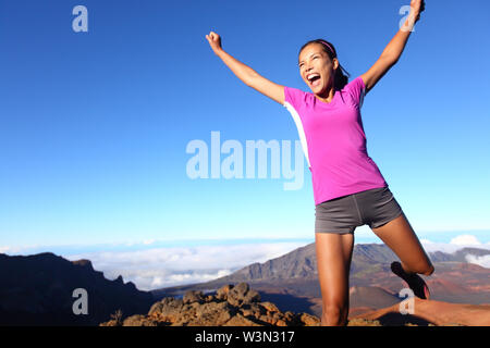 Successo vincitore fitness runner donna jumping happy eccitati ed energici con il tifo felice espressione faccia celebrare. Esecuzione sportiva ragazza tifo dopo la formazione outdoor nel paesaggio di Vulcano. Foto Stock