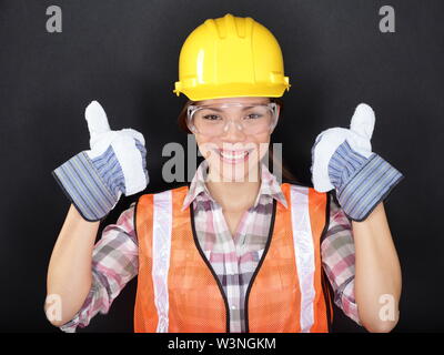 Operaio edile donna felice facendo pollice in alto con protezione usura. Giovane donna che indossa gli occhiali di protezione, giubbotto e Casco giallo per la sicurezza e la protezione su sfondo nero. Asian modello femminile. Foto Stock