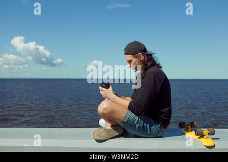 Un giovane uomo in un berretto da baseball con uno smartphone e un giallo skateboard siede sul parapetto della città terrapieno su una soleggiata giornata estiva. Foto Stock