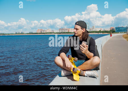 Un giovane uomo in un berretto da baseball con uno smartphone e un giallo skateboard siede sul parapetto della città terrapieno su una soleggiata giornata estiva. Foto Stock