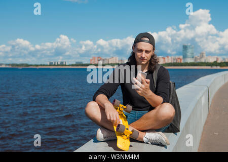 Un giovane uomo in un berretto da baseball con uno smartphone e un giallo skateboard siede sul parapetto della città terrapieno su una soleggiata giornata estiva. Foto Stock