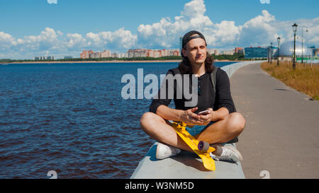 Un giovane uomo in un berretto da baseball con uno smartphone e un giallo skateboard siede sul parapetto della città terrapieno su una soleggiata giornata estiva. Foto Stock