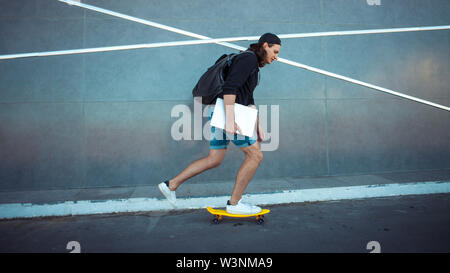 Un giovane uomo in un cappello da baseball, con uno zaino e gli shorts in denim con un laptop sotto il suo braccio, unità passato grigio di una parete di granito con strisce bianche. Foto Stock