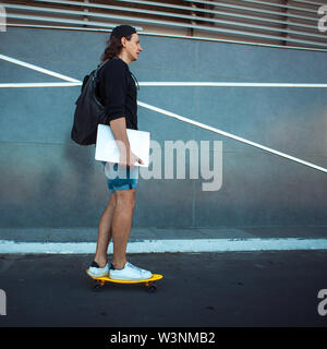 Un giovane uomo in un cappello da baseball, con uno zaino e gli shorts in denim con un laptop sotto il suo braccio, unità passato grigio di una parete di granito con strisce bianche. Foto Stock