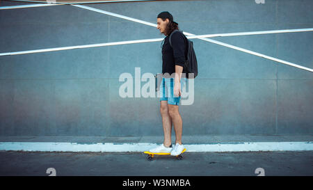 Un giovane uomo in un cappello da baseball, con uno zaino e gli shorts in denim con un laptop sotto il suo braccio, unità passato grigio di una parete di granito con strisce bianche. Foto Stock