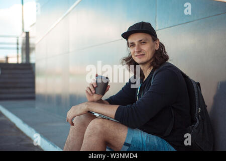 Un giovane uomo in un cappello da baseball, con uno zaino e gli shorts in denim, beve caffè seduti su asfalto, accanto al grigio di una parete di granito. Foto Stock