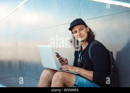 Un giovane uomo, seduto su un giallo sullo skateboard, sta cercando di sincronizzare un computer portatile con un telefono. A quanto pare è accaduto. Egli ha felicemente guardare dentro il telaio. Foto Stock