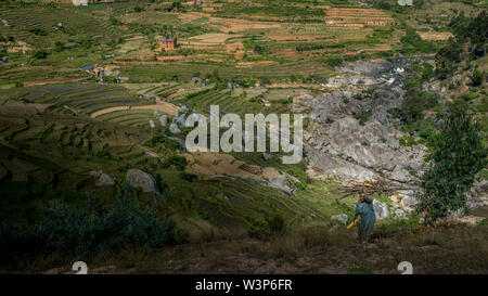 Ricefields e tradizionale Betsileo case sul Madagascar highlands, vicino ad Ambositra, sulla principale N7 road. Old Lady trasportare il legno sul suo capo. Foto Stock