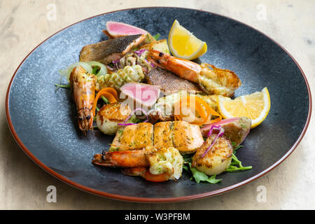 Close up mix di pesce alla griglia e di pesce piatto con insalata fresca, gamberi e capesante, tonno, salmone, spigola, limone, salsa verde in una piastra nera professare Foto Stock