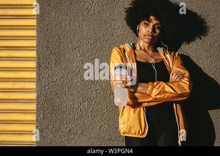 African American donna in usura fitness prendendo una pausa da allenamento in piedi con una bottiglia di acqua in mano. Athletic donna prendendo una pausa durante l'allenamento. Foto Stock