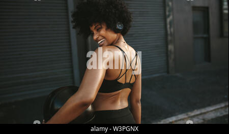 Vista posteriore della donna sorridente in usura fitness all'aperto permanente con la palla medica. Donna africana ascoltando musica su cuffie e riposo dopo wo Foto Stock