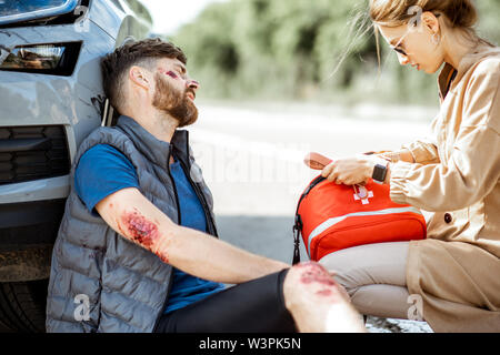 Autista fretta con kit di primo soccorso per aiutare uomo ferito con ferite sanguinanti seduta vicino la vettura dopo l'incidente stradale Foto Stock