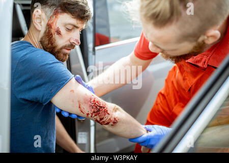 Lavoratore di ambulanza esaminando profonde ferite del braccio di un uomo seduto vicino la vettura dopo l'incidente stradale, fornendo assistenza medica d'emergenza Foto Stock