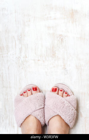Gambe femmina rosso con chiodi in home pellicce soffici pantofole rosa su una luce sullo sfondo di legno. flat laici. Vista dall'alto. Il concetto di un accogliente luminosa casa ragazza. Foto Stock