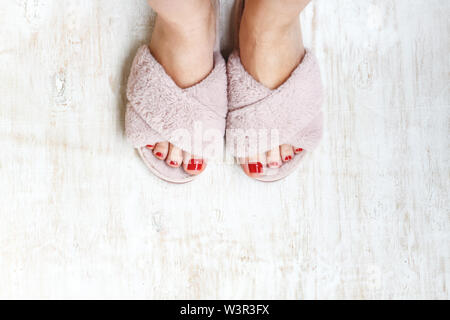 Gambe femmina rosso con chiodi in home pellicce soffici pantofole rosa su una luce sullo sfondo di legno. flat laici. Vista dall'alto. Il concetto di un accogliente luminosa casa ragazza. Foto Stock