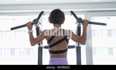 Slim donna sportivo sta facendo pull-ups sulla barra orizzontale con ampia impugnatura in palestra. Ella è in abbigliamento sportivo di fronte alla finestra, vista posteriore. Foto Stock