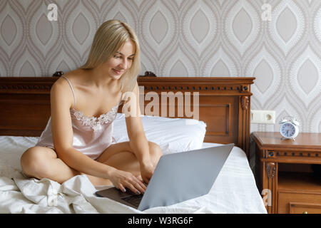 Una giovane bella ragazza bionda si sveglia la mattina e si siede sul letto e si lavora per un laptop. La ragazza si sveglia la mattina Foto Stock