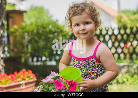 Piccola e dolce ragazza con i capelli ricci sorrisi mentre irrigazione fiori colorati nel bel giardino Foto Stock