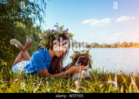 Pug cane e il suo padrone la refrigerazione dal lago indossando ghirlande di fiori. Happy puppy e donna godendo estate natura all'aperto Foto Stock