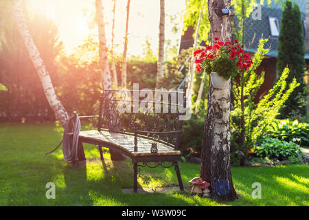 Casa giardino nel cortile con panca, appeso fioritura fiori di tubo flessibile di irrigazione anf alberi. Caldo in estate o in autunno tramonto del tempo. Foto Stock