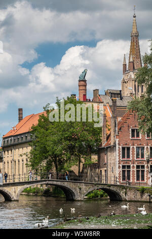 Bruges, Fiandre, Belgio - 17 Giugno 2019: Ponte sul canal con cigni al beghinaggio con birreria e costruzione di camini in retro, plus guglia di Notre Foto Stock