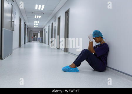 Teso chirurgo femmina con le mani sul fronte seduto sul pavimento del corridoio all'ospedale Foto Stock