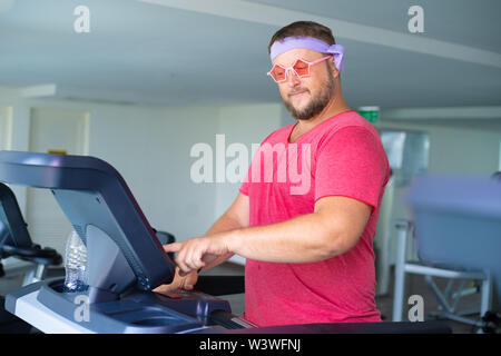 Funny fat maschio in rosa bicchieri e in una t-shirt rosa è impegnato su un tapis roulant in palestra raffigurante una ragazza. l uomo preme un pulsante su un tapis roulant Foto Stock