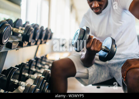 Giovane americano africano uomo seduto e il sollevamento di un manubrio vicino al rack in palestra. Peso maschio persona di formazione facendo un bicipite curl nel centro fitness Foto Stock