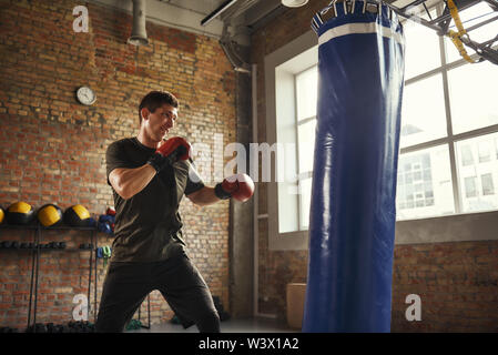 Forte e motivato. Giovane e sportivo boxe uomo in guanti in stile loft palestra. Boxer. Lo sport professionistico. Sacco da boxe Foto Stock