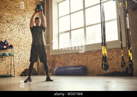 Facendo corpo perfetto. Lunghezza intera di belli e forti uomo con grande nero dumbbell sotto la sua testa mentre ti alleni in palestra. Esercizi di peso. Concetto di bodybuilding. Allenamento. Foto Stock