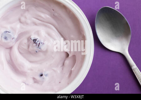 Mirtillo yogurt in una tazza di plastica su sfondo viola con cucchiaio in metallo - vista superiore close up foto di yogurt in contenitore di plastica Foto Stock