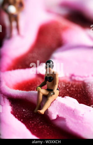 Primo piano di una donna in miniatura che indossa un bikini, occhiali da nuoto e tappo su un gelato alla fragola e un uomo in miniatura con una palla in mano nei Foto Stock