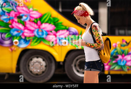 Una donna bionda con un telefono in mano è in piedi contro lo sfondo di un autobus gialli. Ragazza moderna indossa un bendaggio rosso, bracci tatuati. Foto Stock