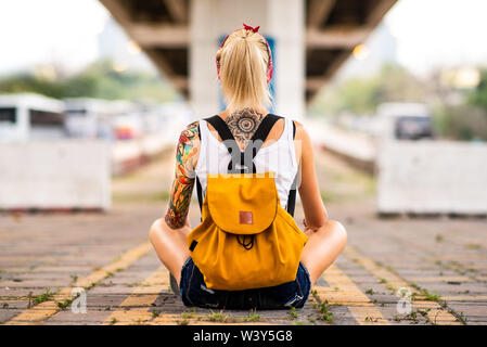 Ragazza moderna si siede con la schiena per la telecamera su una strada di marcatura. Porta un zaino giallo, bracci tatuati Foto Stock